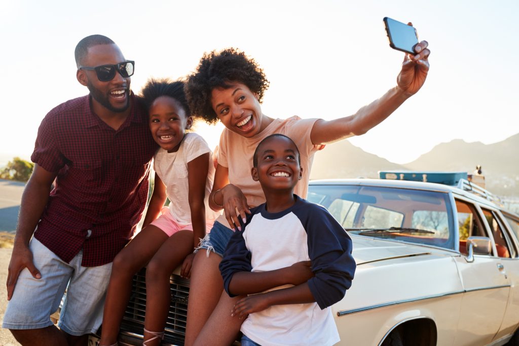 A family with 2 children smile for the selfie while leaning on the hood of a white vehicle with low mountains in the background - on a vacation road trip